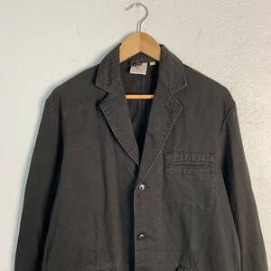 Puede incluir: Blazer de color carb&oacute;n oscuro con solapa de muesca, cierre de tres botones y dos bolsillos delanteros. La chaqueta est&aacute; colgada de una percha de madera contra una pared blanca lisa. El blazer tiene un dise&ntilde;o cl&aacute;sico y a medida.