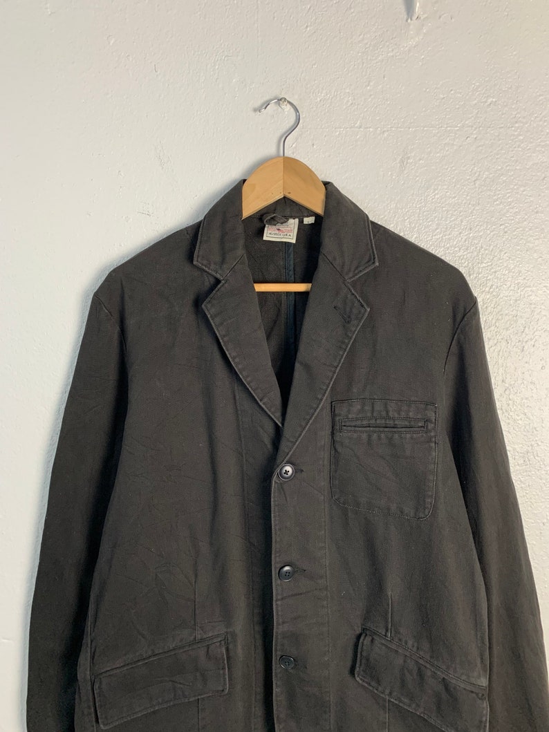 Puede incluir: Blazer de color carb&oacute;n oscuro con solapa de muesca, cierre de tres botones y dos bolsillos delanteros. La chaqueta est&aacute; colgada de una percha de madera contra una pared blanca lisa. El blazer tiene un dise&ntilde;o cl&aacute;sico y a medida.