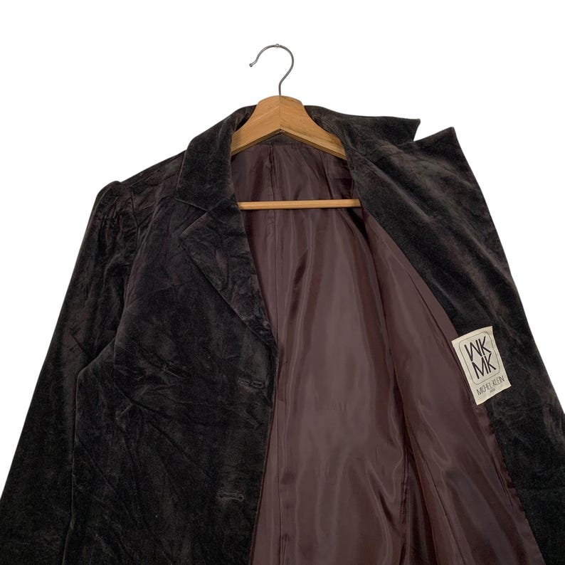 Peut inclure: Un blazer en velours marron fonc&eacute; avec un col crant&eacute; et deux boutons, suspendu &agrave; un cintre en bois. La doublure int&eacute;rieure est de couleur marron clair. Une &eacute;tiquette avec le texte "WK MK MODEL RIN" est visible.