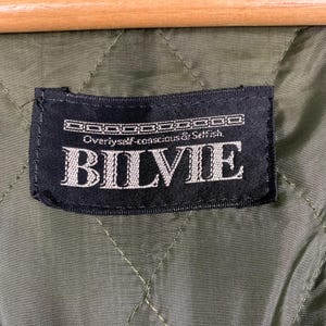Op de afbeelding: Een zwart label met witte tekst dat "Overly self-conscious & Selfish. Bilvie." luidt. Het label is bevestigd aan een groene gewatteerde stof.