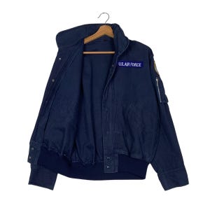 May include: A navy blue bomber jacket with a high collar and a zip pocket on the left sleeve. The jacket features a patch that reads "U.S. AIR FORCE". The jacket is open, revealing the interior lining and a wooden hanger.