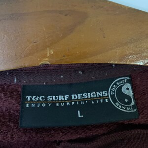 Puede incluir: Una etiqueta en blanco y negro con el texto "T&C Surf Designs Enjoy Surfin' Life" y un s&iacute;mbolo yin yang en blanco y negro con el texto "T&C Surf Hawaii". La etiqueta est&aacute; unida a una sudadera color marr&oacute;n.