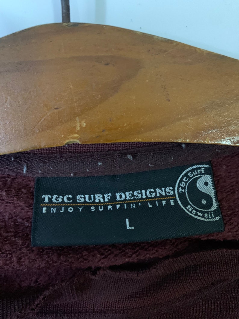 Puede incluir: Una etiqueta en blanco y negro con el texto "T&C Surf Designs Enjoy Surfin' Life" y un s&iacute;mbolo yin yang en blanco y negro con el texto "T&C Surf Hawaii". La etiqueta est&aacute; unida a una sudadera color marr&oacute;n.