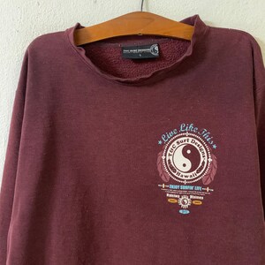 Puede incluir: Una sudadera color marr&oacute;n con un estampado gr&aacute;fico de un s&iacute;mbolo yin yang dentro de un c&iacute;rculo con el texto "Live Like This" y "T&C Surf Designs Hawaii" alrededor. El gr&aacute;fico tambi&eacute;n incluye el texto "Enjoy Surfin' Life" y los nombres de dos lugares, "Haleiwa" y "Waimea".