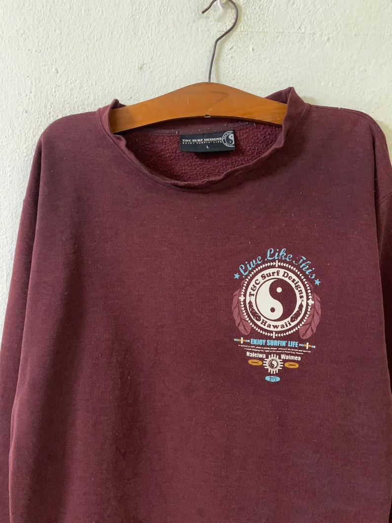 Puede incluir: Una sudadera color marr&oacute;n con un estampado gr&aacute;fico de un s&iacute;mbolo yin yang dentro de un c&iacute;rculo con el texto "Live Like This" y "T&C Surf Designs Hawaii" alrededor. El gr&aacute;fico tambi&eacute;n incluye el texto "Enjoy Surfin' Life" y los nombres de dos lugares, "Haleiwa" y "Waimea".