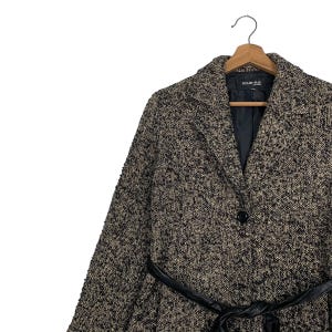 Puede incluir: Un abrigo de tweed en tonos negros, marrones y beige, con un cintur&oacute;n de cuero negro. El abrigo tiene un cuello con solapas, un cierre de un solo bot&oacute;n y la marca "noue-rue" es visible en la etiqueta.