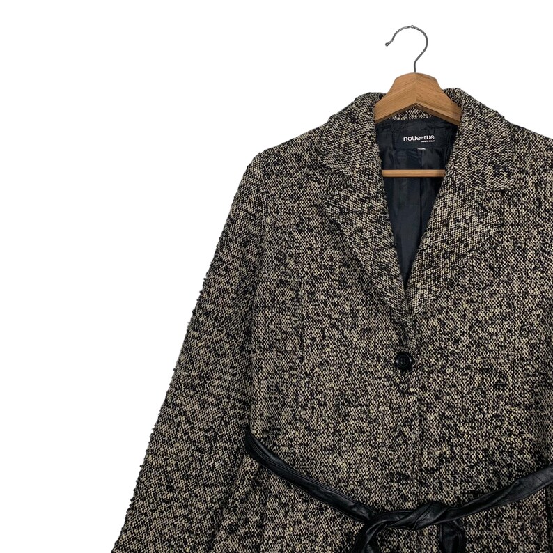 Puede incluir: Un abrigo de tweed en tonos negros, marrones y beige, con un cintur&oacute;n de cuero negro. El abrigo tiene un cuello con solapas, un cierre de un solo bot&oacute;n y la marca "noue-rue" es visible en la etiqueta.