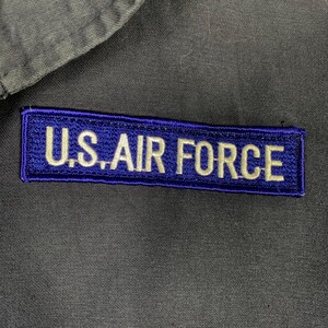 May include: A rectangular patch with the text "U.S. AIR FORCE" embroidered in white on a dark blue background. The patch is sewn onto a gray fabric, possibly a uniform.