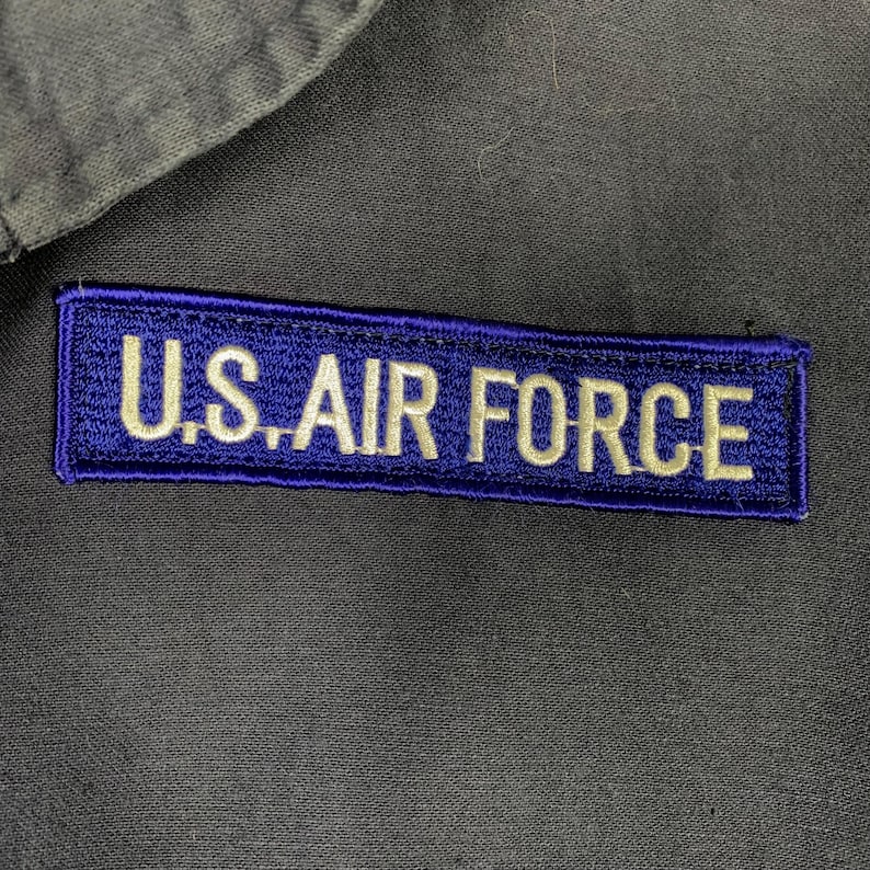 May include: A rectangular patch with the text "U.S. AIR FORCE" embroidered in white on a dark blue background. The patch is sewn onto a gray fabric, possibly a uniform.