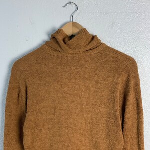 Puede incluir: Un su&eacute;ter de cuello alto marr&oacute;n con una textura suave y peluda. El su&eacute;ter est&aacute; colgado en una percha de madera.