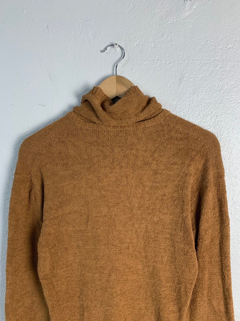 Puede incluir: Un su&eacute;ter de cuello alto marr&oacute;n con una textura suave y peluda. El su&eacute;ter est&aacute; colgado en una percha de madera.