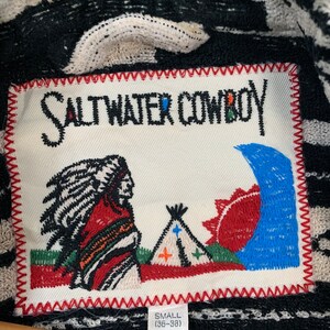 Puede incluir: Un parche blanco con texto bordado en negro que dice "Saltwater Cowboy" con una imagen bordada en rojo, blanco y azul de un hombre nativo americano, una tienda de campa&ntilde;a y un sol. El parche tiene un borde cosido rojo y el texto "Small (36-38)" en negro.