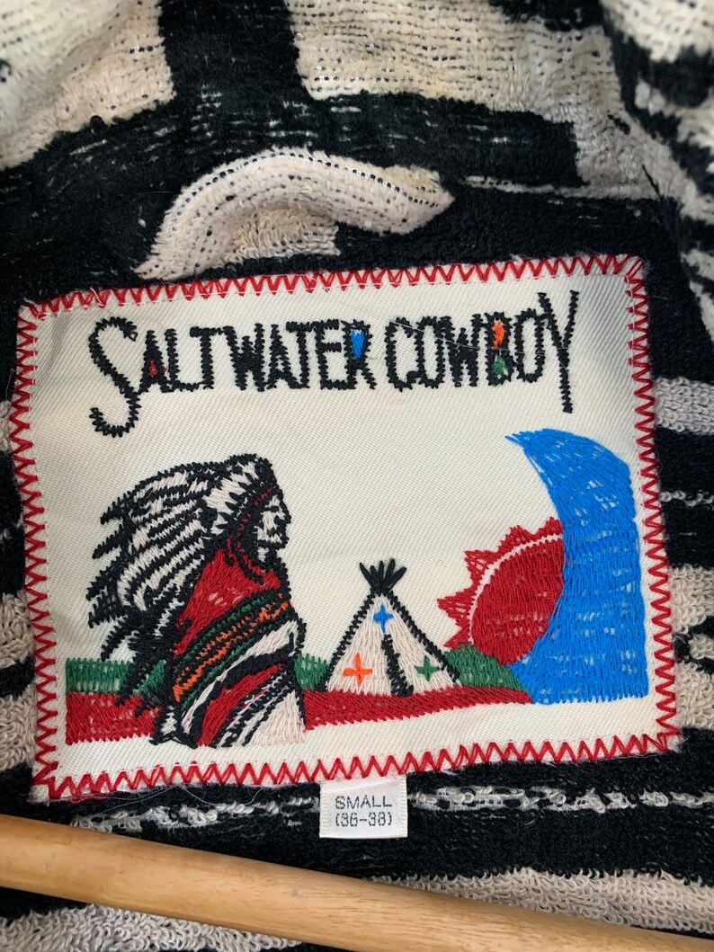 Puede incluir: Un parche blanco con texto bordado en negro que dice "Saltwater Cowboy" con una imagen bordada en rojo, blanco y azul de un hombre nativo americano, una tienda de campa&ntilde;a y un sol. El parche tiene un borde cosido rojo y el texto "Small (36-38)" en negro.