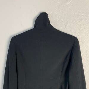 May include: A black blazer with a mock neck collar. The blazer is hanging on a white wall.