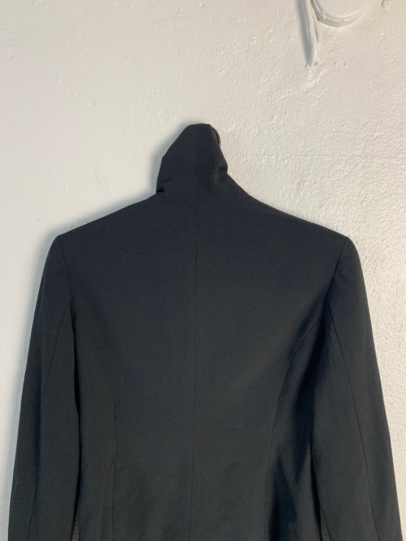 May include: A black blazer with a mock neck collar. The blazer is hanging on a white wall.