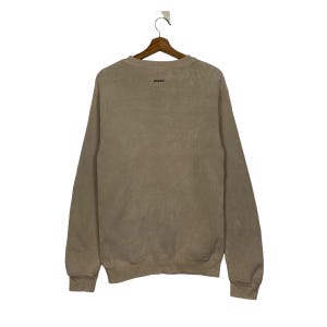 May include: A light brown sweatshirt with a crew neck and long sleeves. The sweatshirt has a small, black logo on the front.