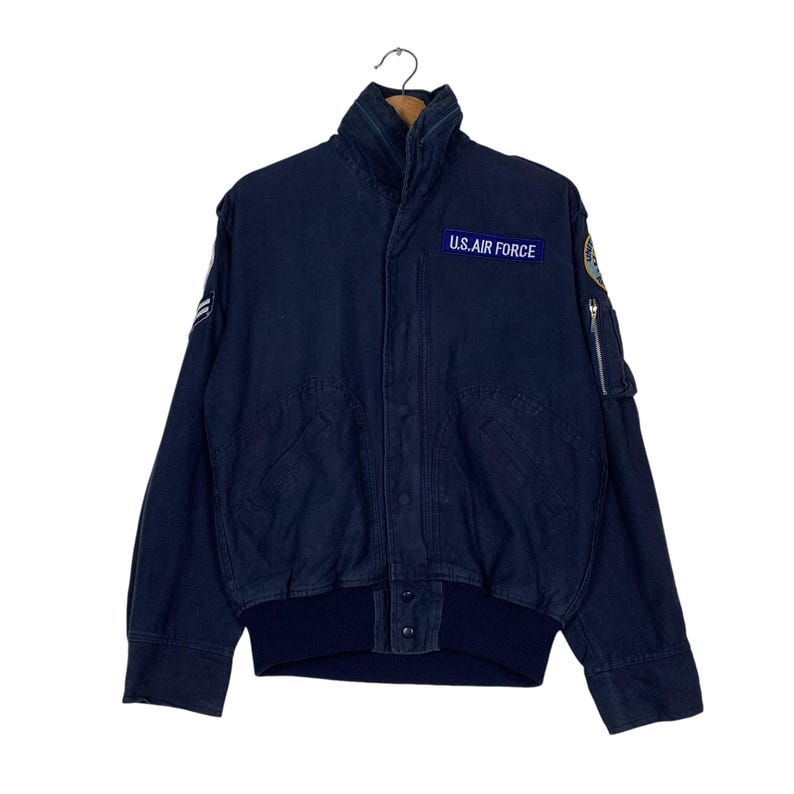 May include: A navy blue bomber jacket with a high collar and a ribbed hem. The jacket features a patch that reads "U.S. AIR FORCE" and a small zippered pocket on the sleeve. The jacket is hanging on a wooden hanger.