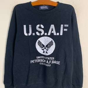 Könnte beinhalten: Schwarzer Pullover mit einem weißen Aufdruck von "U.S.A.F." und einem militärischen Abzeichen. Der Text unter dem Abzeichen lautet "United States Peterson A.F. Base Colorado."