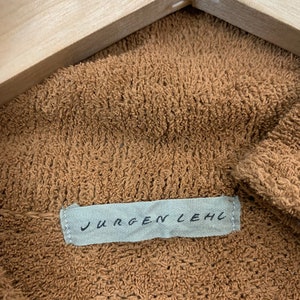 Puede incluir: Una tela de punto marr&oacute;n, texturizada, con una etiqueta blanca que dice "JURGEN LEHL" en letras negras.