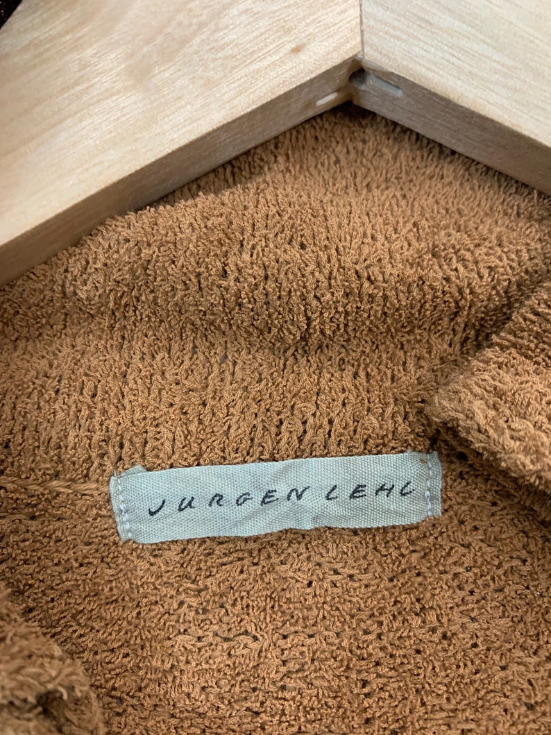 Puede incluir: Una tela de punto marr&oacute;n, texturizada, con una etiqueta blanca que dice "JURGEN LEHL" en letras negras.