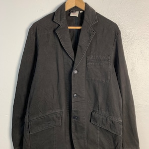 Puede incluir: Blazer marr&oacute;n oscuro con cierre de tres botones, dos bolsillos delanteros y un bolsillo en el pecho. La chaqueta est&aacute; colgada de una percha de madera. El blazer tiene un dise&ntilde;o cl&aacute;sico y a medida.