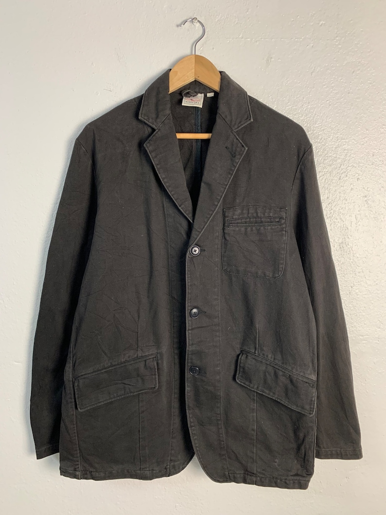 Puede incluir: Blazer marr&oacute;n oscuro con cierre de tres botones, dos bolsillos delanteros y un bolsillo en el pecho. La chaqueta est&aacute; colgada de una percha de madera. El blazer tiene un dise&ntilde;o cl&aacute;sico y a medida.