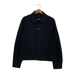 Può includere: Giacca nera con bottoni e colletto. La giacca ha maniche lunghe e due tasche anteriori. È appesa a un'appendiabiti di legno.