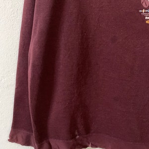 Puede incluir: Una camisa de manga larga color granate con un dise&ntilde;o gr&aacute;fico en la parte delantera. El dise&ntilde;o presenta un tocado nativo americano con plumas y cuentas.
