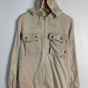 May include: A beige linen jacket with a hood and a zipper closure. The jacket has two large pockets on the front and a drawstring at the waist.