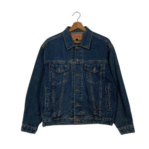 Op de afbeelding: Een donkerblauw denim jack met een knoopsluiting en twee voorzakken. Het jack heeft een rood label met de tekst "TOPSHOP" aan de linkerkant van de borst.