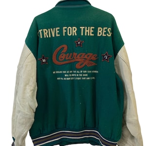 Könnte beinhalten: Grün-weiße Baseballjacke mit einem großen gestickten "Courage"-Logo auf der Rückseite. Die Jacke hat einen weißen Lederärmel und einen grünen Wollkörper. Der Text "Strive for the Best" ist über dem Logo gestickt. Der Text "We declare that at any time all of our team members will be united in the game and will do our best to fight fair and square" ist unter dem Logo gestickt.