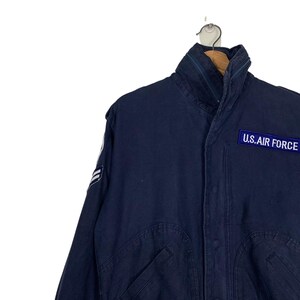 May include: Navy blue jacket with a high collar and the text "U.S. AIR FORCE" on a patch. The jacket has a zipper and a military-style design. The jacket is hanging on a metal hanger.