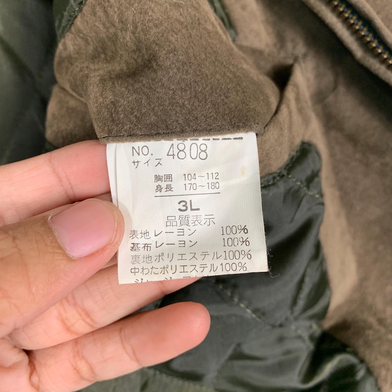 Op de afbeelding: Een groene jas met een label waarop "NO. 4808" staat en met maatgegevens: "Borst 104-112 cm, Lengte 170-180 cm, 3L". Het label vermeldt ook de stofsamenstelling: "Buitenstof: Rayon 100%, Voering: Rayon 100%, Vulling: Polyester 100%, Tussenvoering: Polyester 100%".