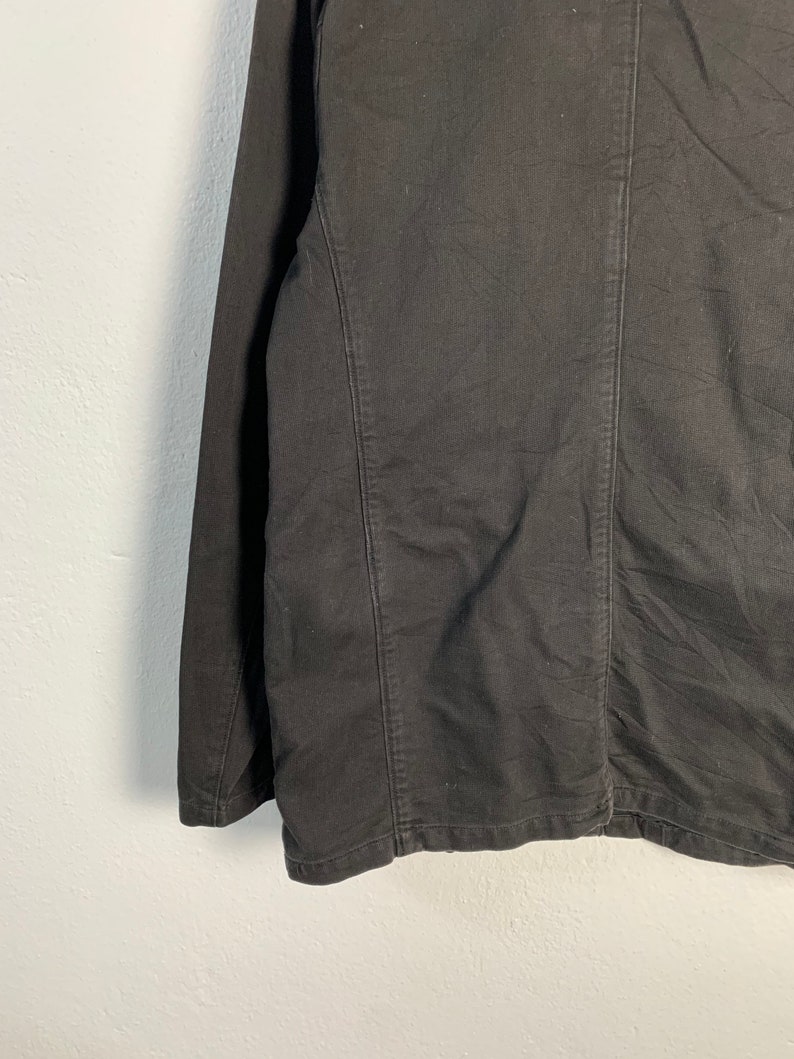 Puede incluir: Primer plano de una chaqueta negra. La chaqueta tiene un dise&ntilde;o a medida con costuras visibles y una silueta estructurada. La tela parece ser un material resistente, posiblemente algod&oacute;n o una mezcla, con una superficie ligeramente texturizada.