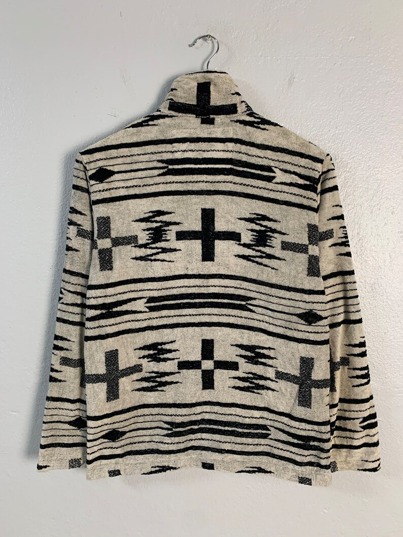 Puede incluir: Un su&eacute;ter de manga larga beige y negro con un patr&oacute;n geom&eacute;trico de cruces negras y l&iacute;neas. El su&eacute;ter tiene un cuello alto y est&aacute; colgado en una percha.