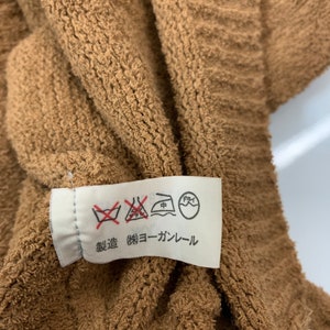 Puede incluir: Una etiqueta de cuidado blanca sobre un fondo marr&oacute;n. La etiqueta tiene varios s&iacute;mbolos, incluyendo una lavadora con una cruz, una plancha con un punto dentro y un c&iacute;rculo con una mano dentro. La etiqueta tambi&eacute;n tiene el texto "無蛍光 (無蛍光) コーラル" en japon&eacute;s.