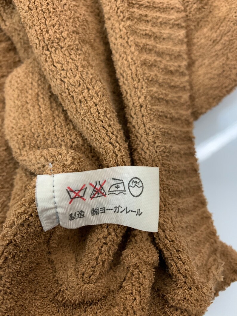 Puede incluir: Una etiqueta de cuidado blanca sobre un fondo marr&oacute;n. La etiqueta tiene varios s&iacute;mbolos, incluyendo una lavadora con una cruz, una plancha con un punto dentro y un c&iacute;rculo con una mano dentro. La etiqueta tambi&eacute;n tiene el texto "無蛍光 (無蛍光) コーラル" en japon&eacute;s.