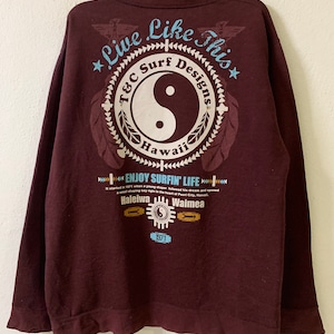 Puede incluir: Una camiseta de manga larga de color marr&oacute;n con un estampado gr&aacute;fico de un s&iacute;mbolo yin-yang dentro de un c&iacute;rculo con el texto "T&C Surf Designs Hawaii" y el texto "Live Like This" encima del c&iacute;rculo. El texto "Enjoy Surfin' Life" est&aacute; impreso debajo del c&iacute;rculo con el texto "Haleiwa" y "Waimea" debajo. El texto "1971" est&aacute; impreso debajo de las palabras "Haleiwa" y "Waimea".