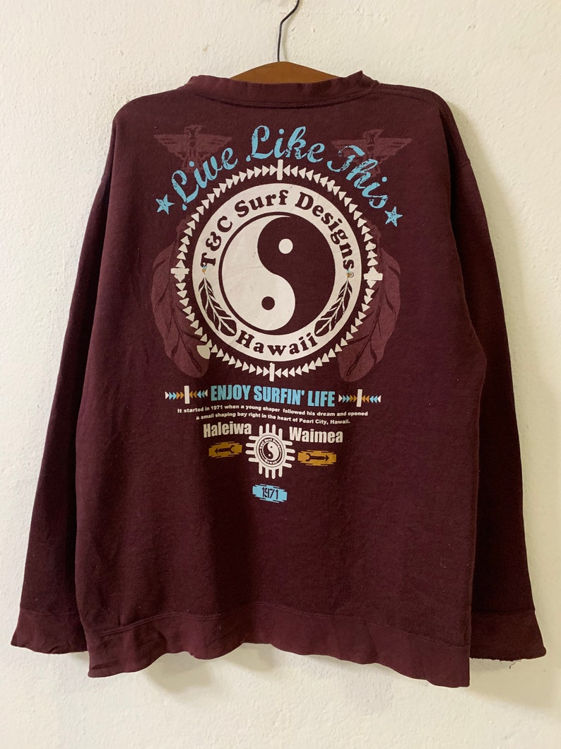 Puede incluir: Una camiseta de manga larga de color marr&oacute;n con un estampado gr&aacute;fico de un s&iacute;mbolo yin-yang dentro de un c&iacute;rculo con el texto "T&C Surf Designs Hawaii" y el texto "Live Like This" encima del c&iacute;rculo. El texto "Enjoy Surfin' Life" est&aacute; impreso debajo del c&iacute;rculo con el texto "Haleiwa" y "Waimea" debajo. El texto "1971" est&aacute; impreso debajo de las palabras "Haleiwa" y "Waimea".