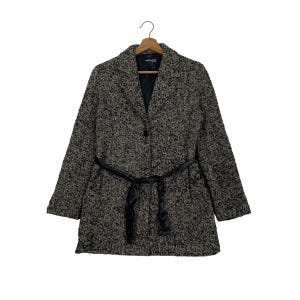 Puede incluir: Un abrigo de tweed negro y beige, con cuello de solapa, mangas largas y cintur&oacute;n negro. El abrigo est&aacute; abotonado y colgado de una percha de madera, mostrando un dise&ntilde;o cl&aacute;sico y a medida.