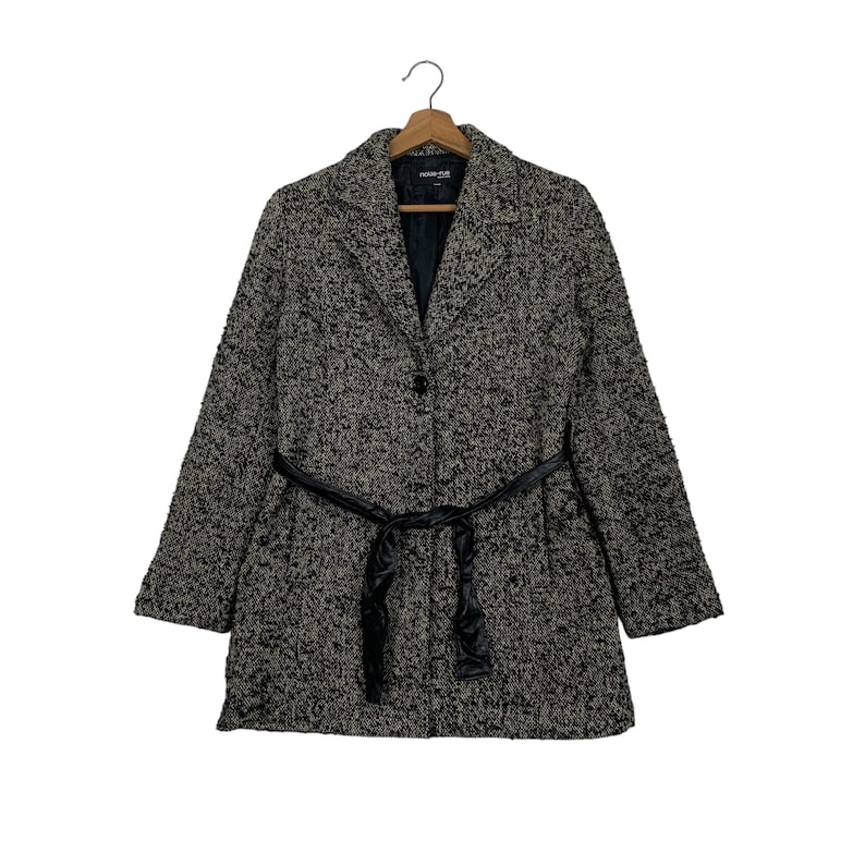 Puede incluir: Un abrigo de tweed negro y beige, con cuello de solapa, mangas largas y cintur&oacute;n negro. El abrigo est&aacute; abotonado y colgado de una percha de madera, mostrando un dise&ntilde;o cl&aacute;sico y a medida.