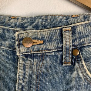 Op de afbeelding: Close-up van de tailleband van een spijkerbroek van blauwe denim met een bruin leren patch waarop "Champion" in witte letters staat.