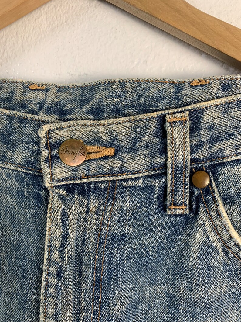 Op de afbeelding: Close-up van de tailleband van een spijkerbroek van blauwe denim met een bruin leren patch waarop "Champion" in witte letters staat.