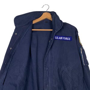 May include: Navy blue U.S. Air Force jacket with a zip closure and a patch on the chest. The jacket is hanging on a wooden hanger. The jacket has a high collar and multiple pockets.