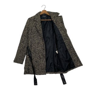 Puede incluir: Un abrigo de estilo tweed en tonos marrones y negros, colgado de una percha de madera. El abrigo presenta un cuello con muescas, un forro de sat&eacute;n negro y un cintur&oacute;n a juego. El abrigo est&aacute; abierto, revelando el forro interior y una etiqueta.