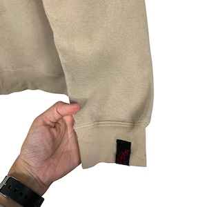 May include: A beige sweatshirt with a black and red logo on the cuff. The logo is a stylized figure with a red outline.