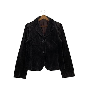 Peut inclure: Un blazer en velours marron fonc&eacute; avec un col classique et une fermeture &agrave; deux boutons. La veste est suspendue &agrave; un cintre en bois sur fond blanc. Le blazer a des manches longues et une coupe ajust&eacute;e.