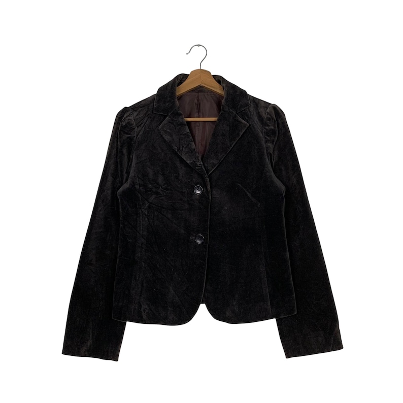 Peut inclure: Un blazer en velours marron fonc&eacute; avec un col classique et une fermeture &agrave; deux boutons. La veste est suspendue &agrave; un cintre en bois sur fond blanc. Le blazer a des manches longues et une coupe ajust&eacute;e.