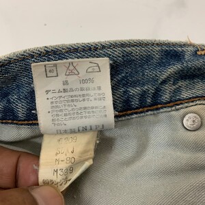 Op de afbeelding: Een close-up van een blauw denim label met witte tekst. Het label zegt "100% Katoen" en bevat wasinstructies. Het label bevat ook de tekst "Verzorgingsinstructies voor denimproducten" en "Gemaakt in Japan". Het label bevat ook de tekst "NIP" en de nummers "6209", "M-80" en "M389".