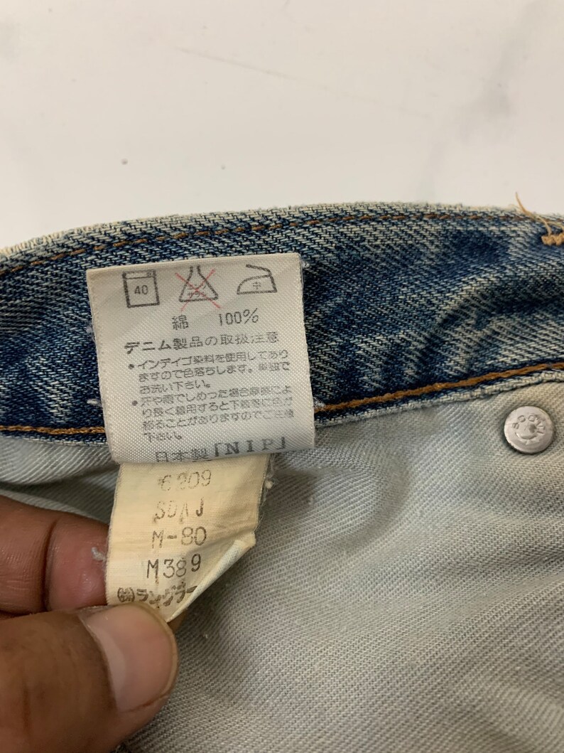 Op de afbeelding: Een close-up van een blauw denim label met witte tekst. Het label zegt "100% Katoen" en bevat wasinstructies. Het label bevat ook de tekst "Verzorgingsinstructies voor denimproducten" en "Gemaakt in Japan". Het label bevat ook de tekst "NIP" en de nummers "6209", "M-80" en "M389".
